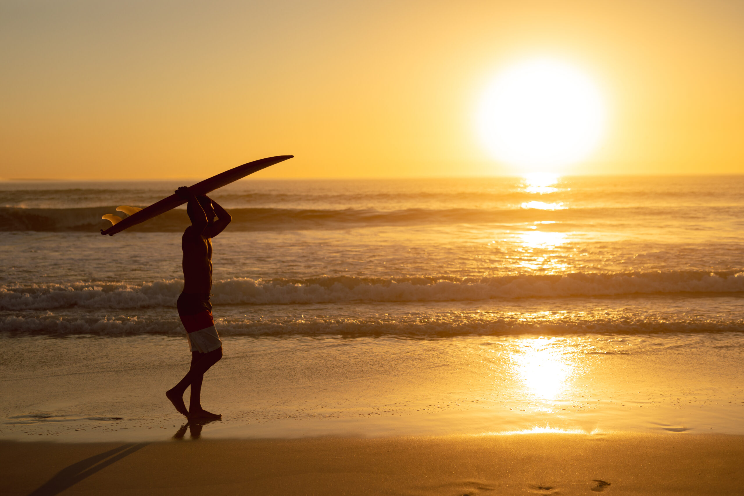 Man walking with surfboard on his head at beach during sunset